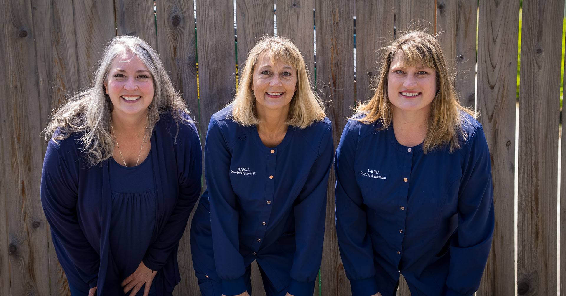 Three smiling Burien dental team members in front of wooden fence