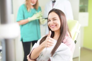 Teenage girl with brown hair sitting in dental chair smiling, winking, and giving a thumbs up